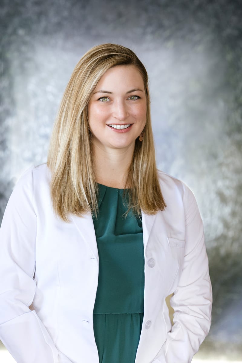 Laura Bolmey, FNP, smiling in a white coat over a green dress, against a grey studio backdrop