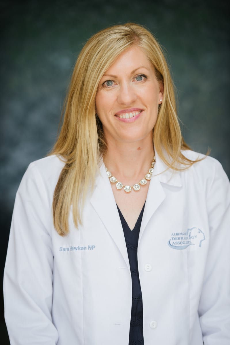 Sara Hawken, NP, smiling in a white coat embroidered with her name and the practice logo, against a dark studio backdrop