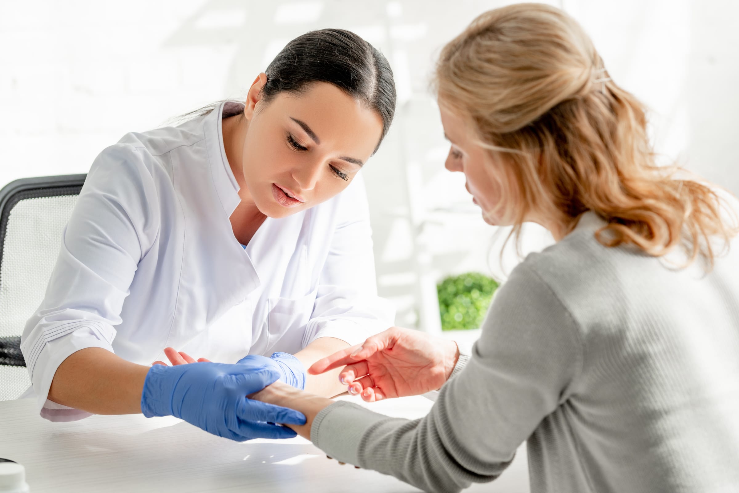 Stock photograph of models: a dermatologist in blue gloves examines the skin of a woman’s forearm across a white desk