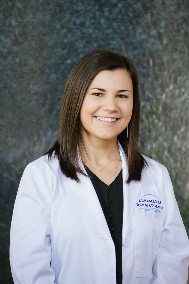 Sara Wilson, PA, smiling in a white coat embroidered with the Albemarle Dermatology Associates logo, against a grey backdrop