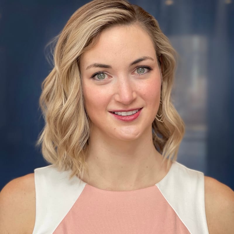 Laura Bolmey, smiling, with wavy shoulder-length blonde hair and a pink and white top, against a dark blue backdrop
