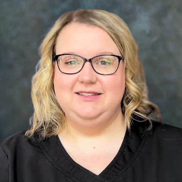 Heather Southers, smiling, in glasses and dark scrubs, against a mottled grey backdrop
