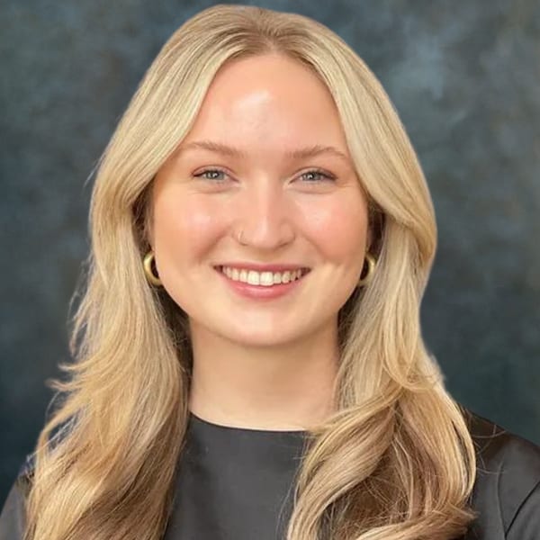 Kalli Dulling, smiling, with long blonde hair and gold hoop earrings, against a mottled grey backdrop