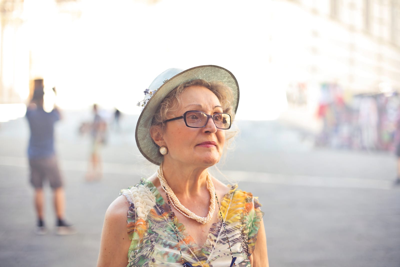 An older woman in a straw sun hat, glasses and earrings, outdoors on a bright, sunny promenade