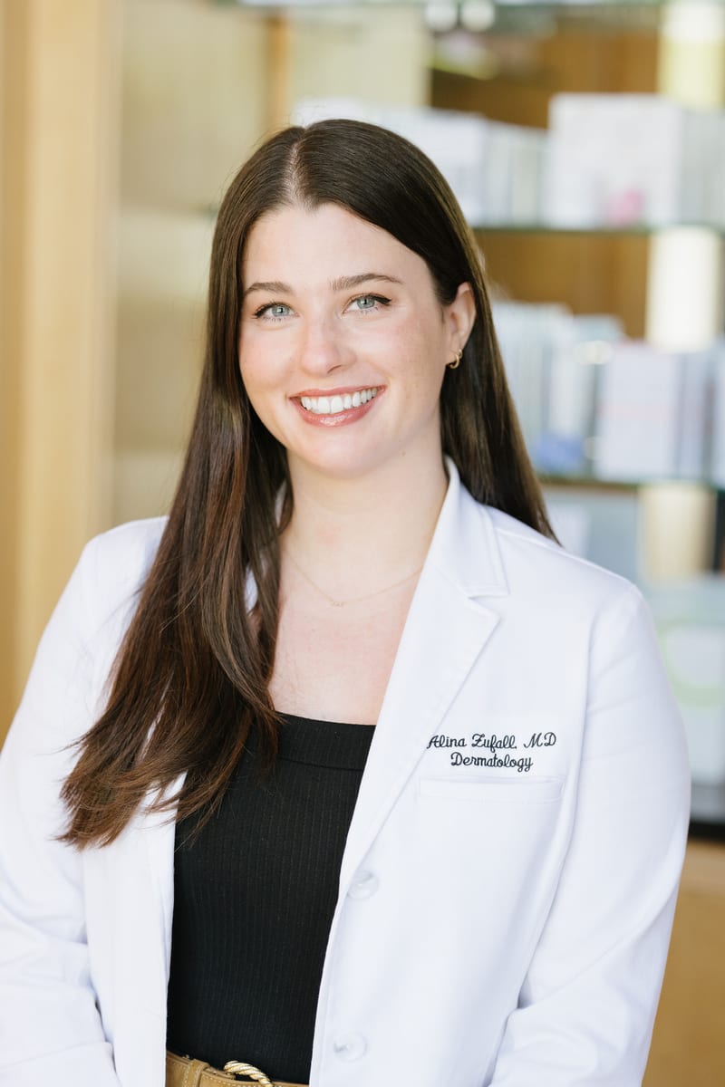 Dr. Alina Zufall smiling in a white coat embroidered with her name, in front of the practice’s product shelves